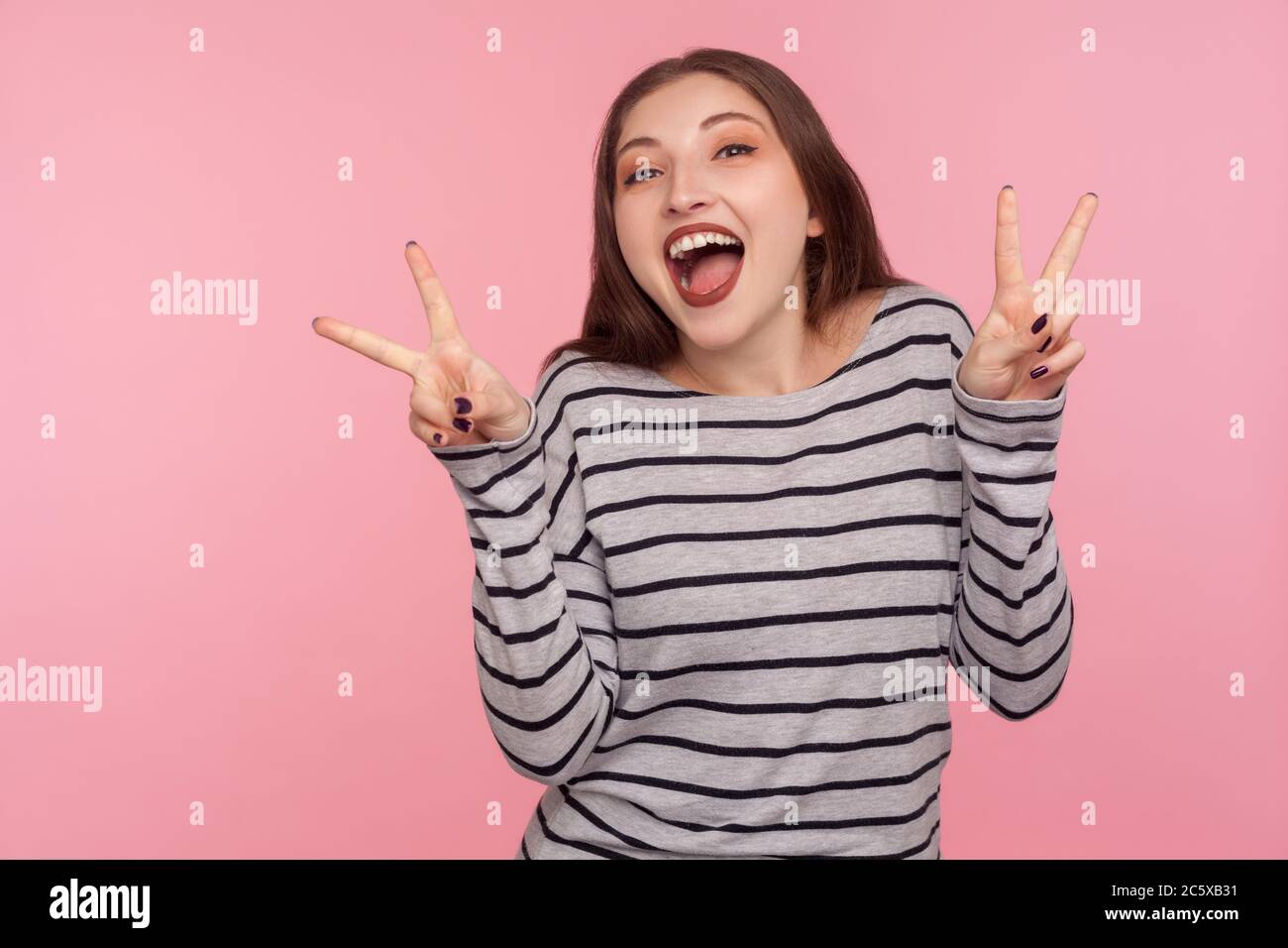 Ritratto di ottimista donna felice in felpa a righe mostrando doppio gesto di vittoria, segno di mano di pace, celebrando la vittoria, felice di successo. ind Foto Stock