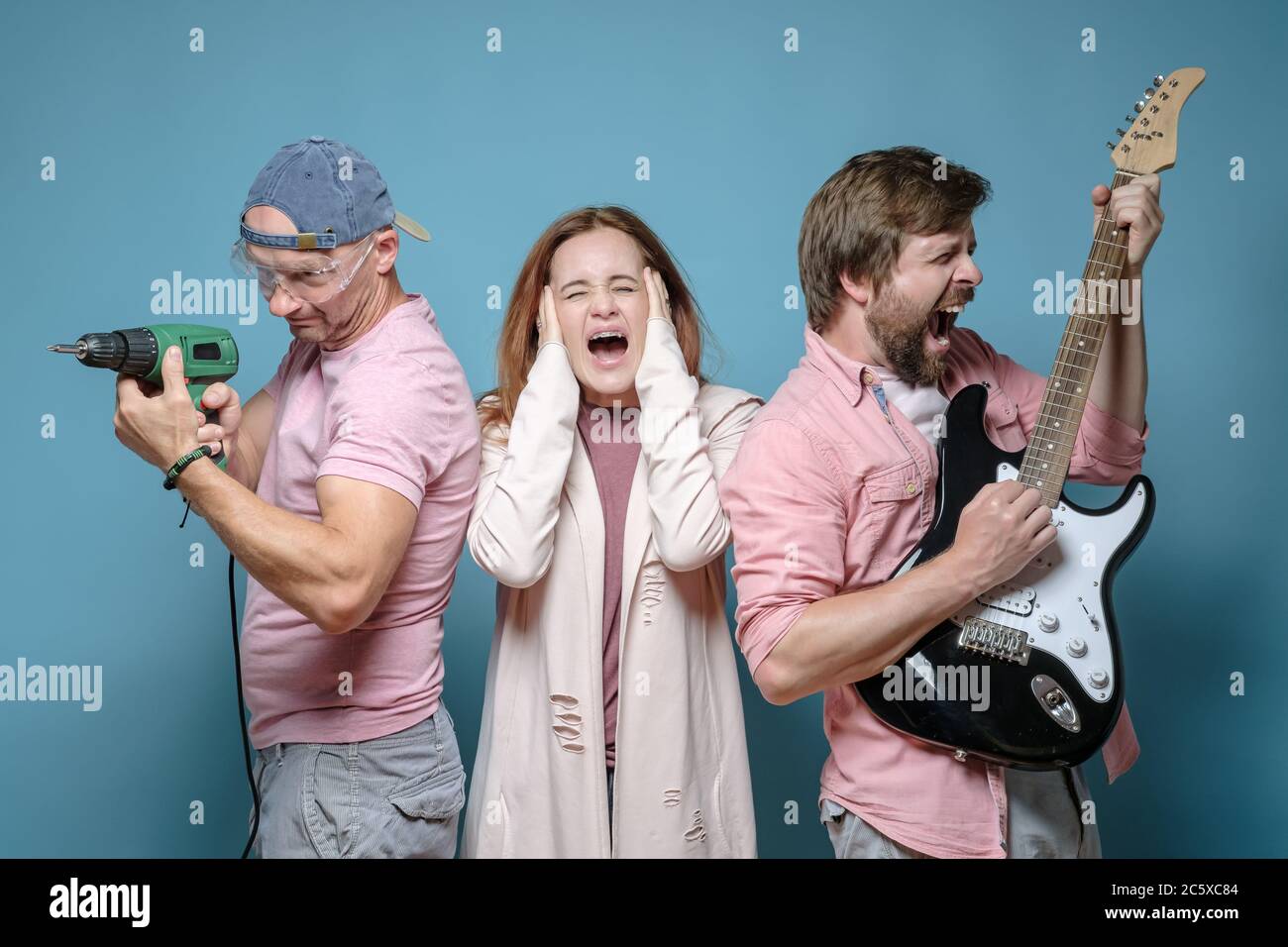Due uomini annoiano una donna, fanno rumore con una chitarra elettrica e un trapano di potenza. Vicino inquieto. Studio, sfondo blu. Foto Stock