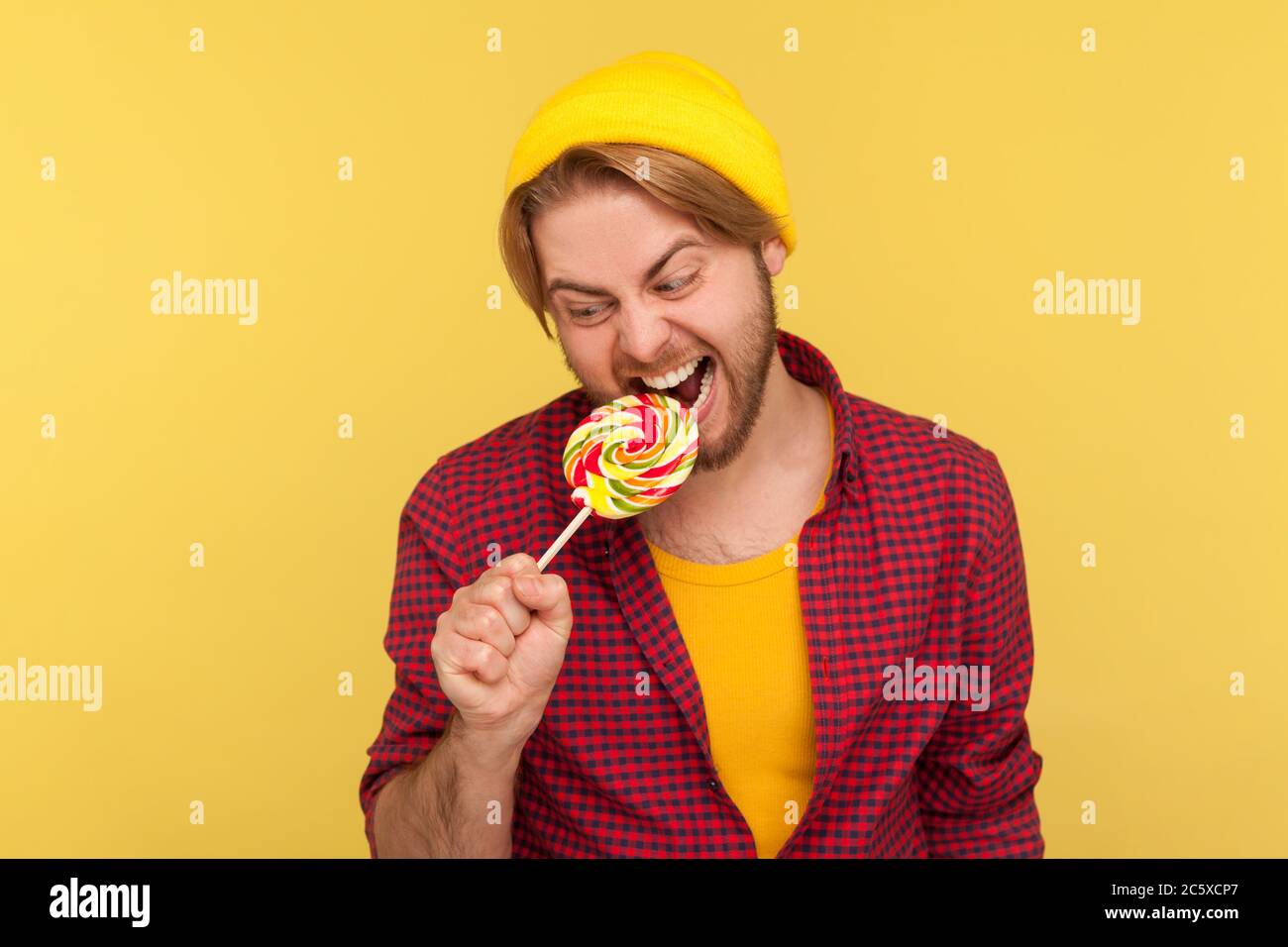 Ragazzo hipster eccitato in cappello di berretto e camicia a scacchi che mordono caramella lollipop, cercando con espressione pazzesca, desiderando mangiare pasticceria dolce con gr Foto Stock