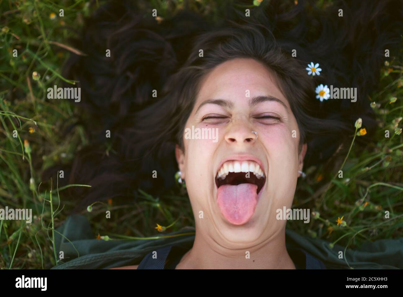 Ritratto di una giovane donna sdraiata nell'erba con fiori nei capelli. Lei attacca la sua lingua con un'espressione felice Foto Stock