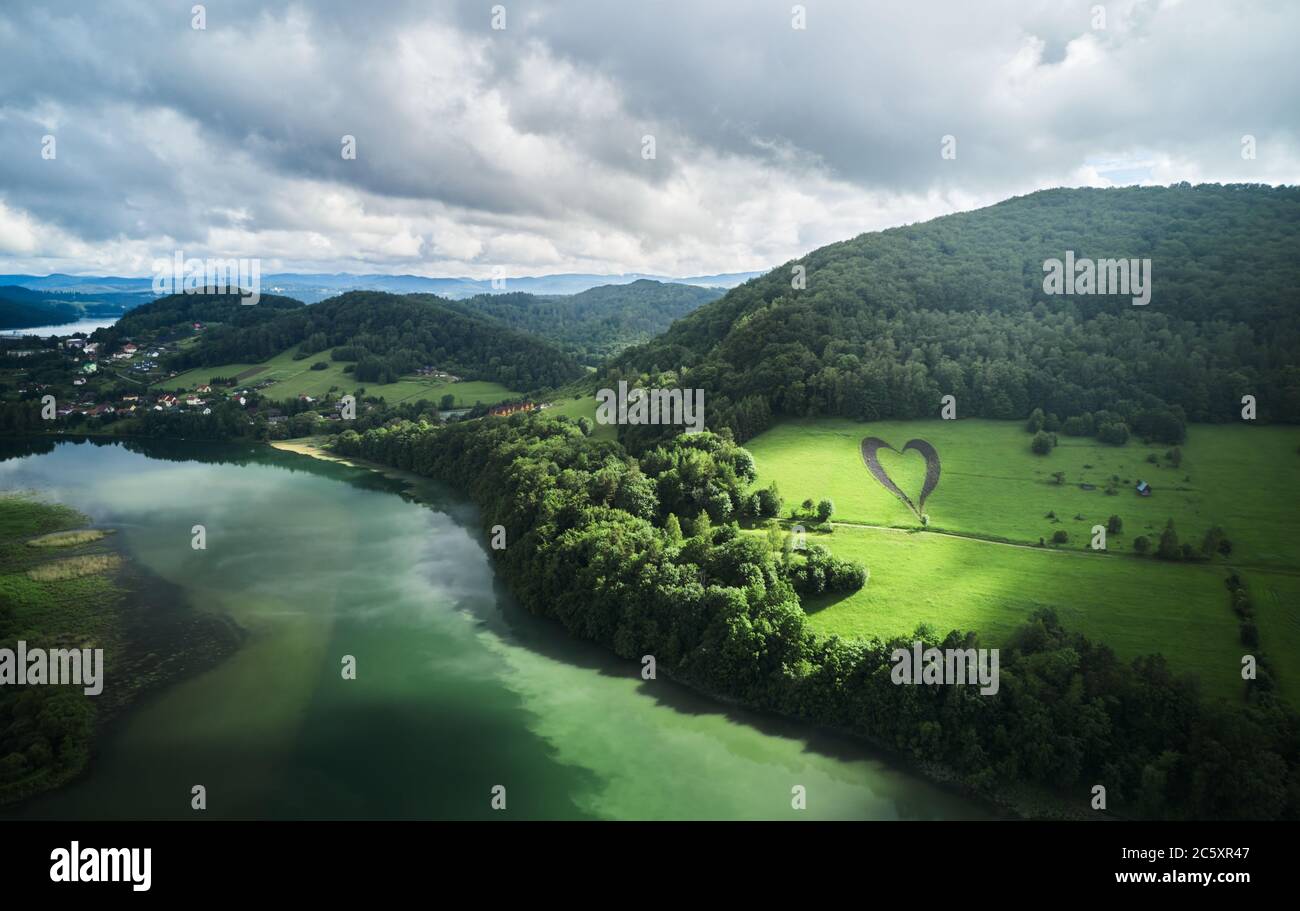 Forma del cuore su una collina verde circondata da montagne, alberi, fiume e solitaria strada vuota. Amore segno in memoria di un amato perso a Myczkowce, Polonia. Foto Stock