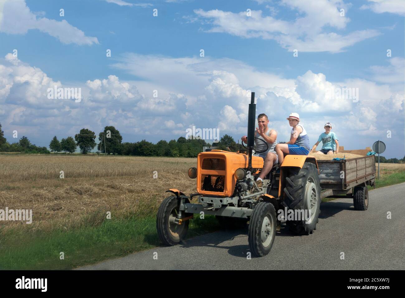 Coltivatore che ondeggiava mentre guidava trattore con moglie e figlia e carro archiviato con grano. Zawady Gmina Rzeczyca Polonia Foto Stock