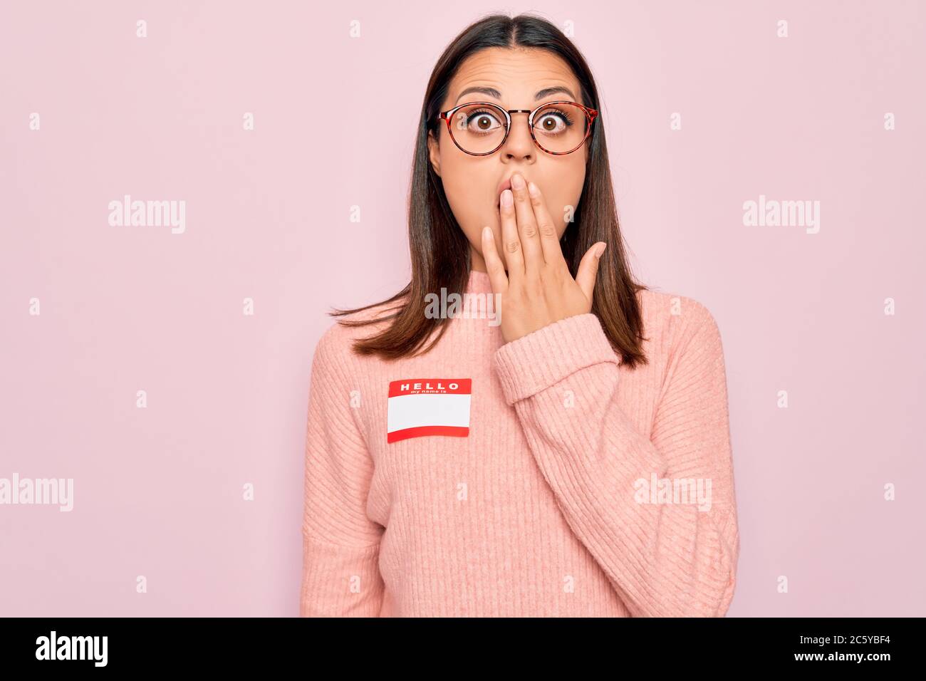 Giovane bella donna bruna con adesivo con ciao il mio nome è messaggio che copre la bocca con la mano, scioccato e impaurito per errore. Sorpresa expre Foto Stock