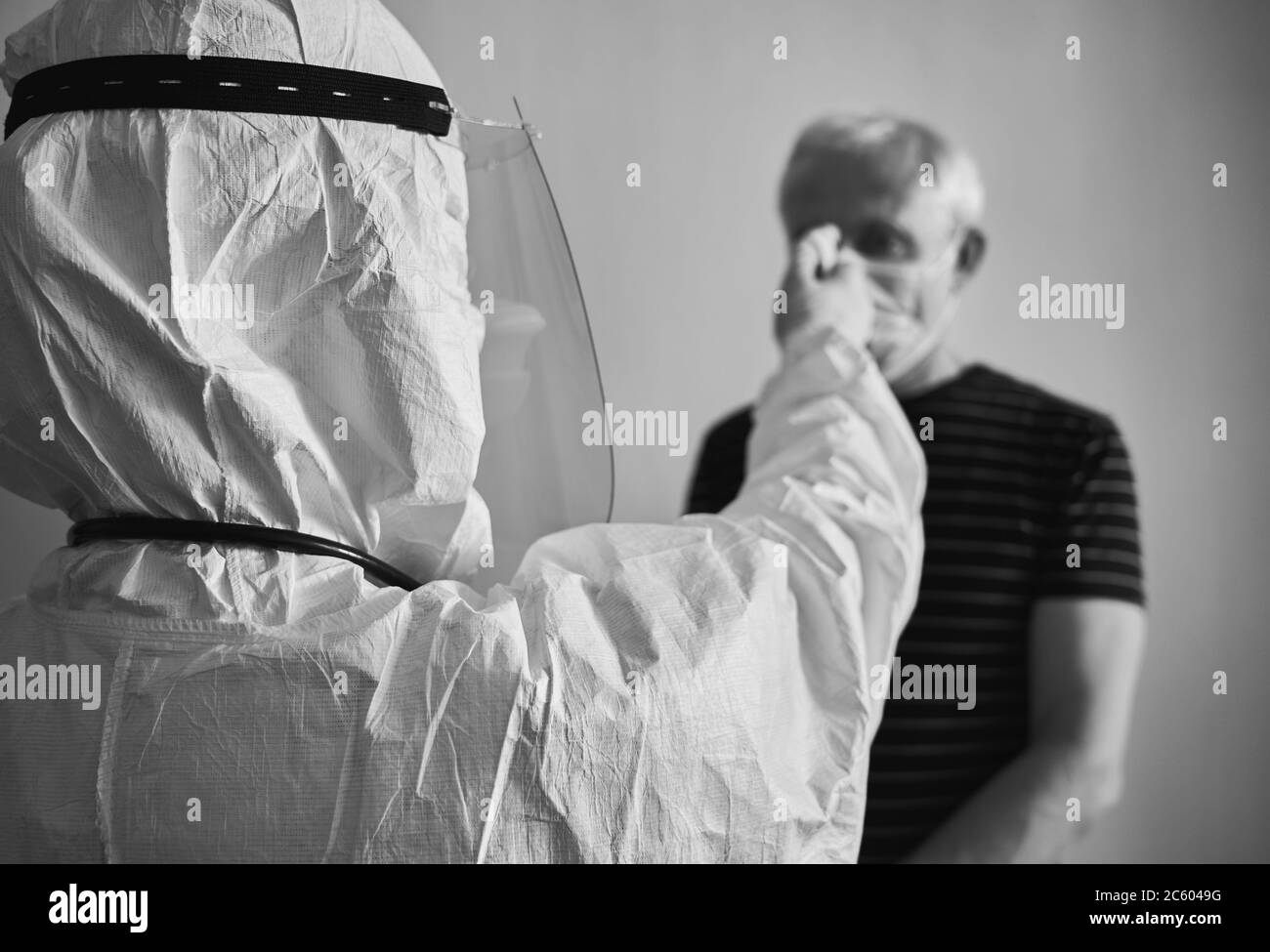 Immagine del medico che misura la febbre dell'uomo anziano con termometro a infrarossi senza contatto. Istantanea bianca e nera della visita del medico a casa del paziente durante la quarantena. Concetto di Covid-19 Foto Stock