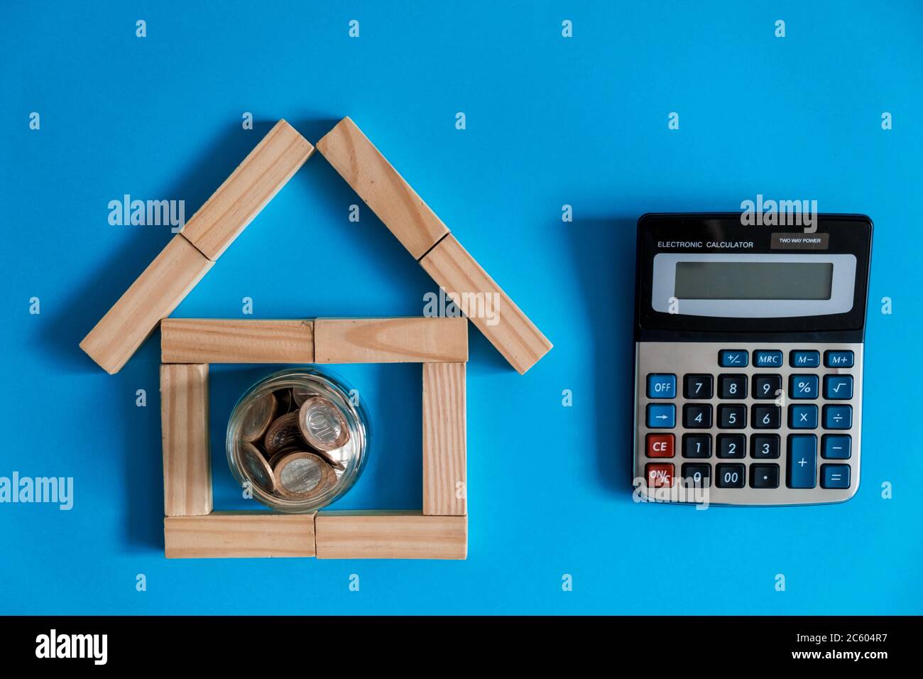 mano che inserisce moneta in legno modello casa su pila di monete su verde erba finta e calcolatrice con gradiente sfondo blu per il risparmio o concetto di investimento domestico Foto Stock