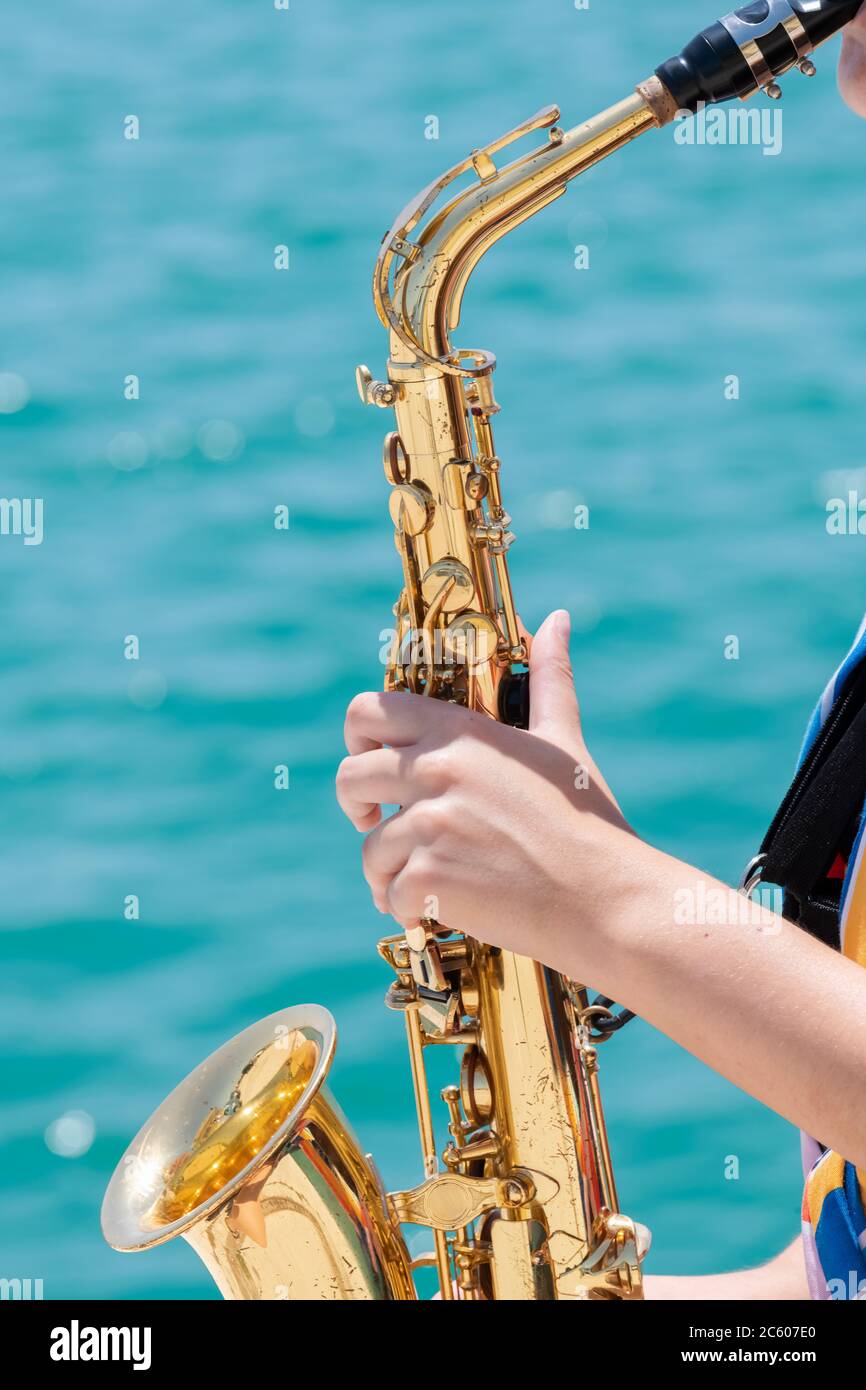 Primo piano di un sassofono dorato brillante tenuto e interpretato da una donna su uno sfondo di mare fuori fuoco. Musicista di strada e concetto di stile di vita. Foto Stock