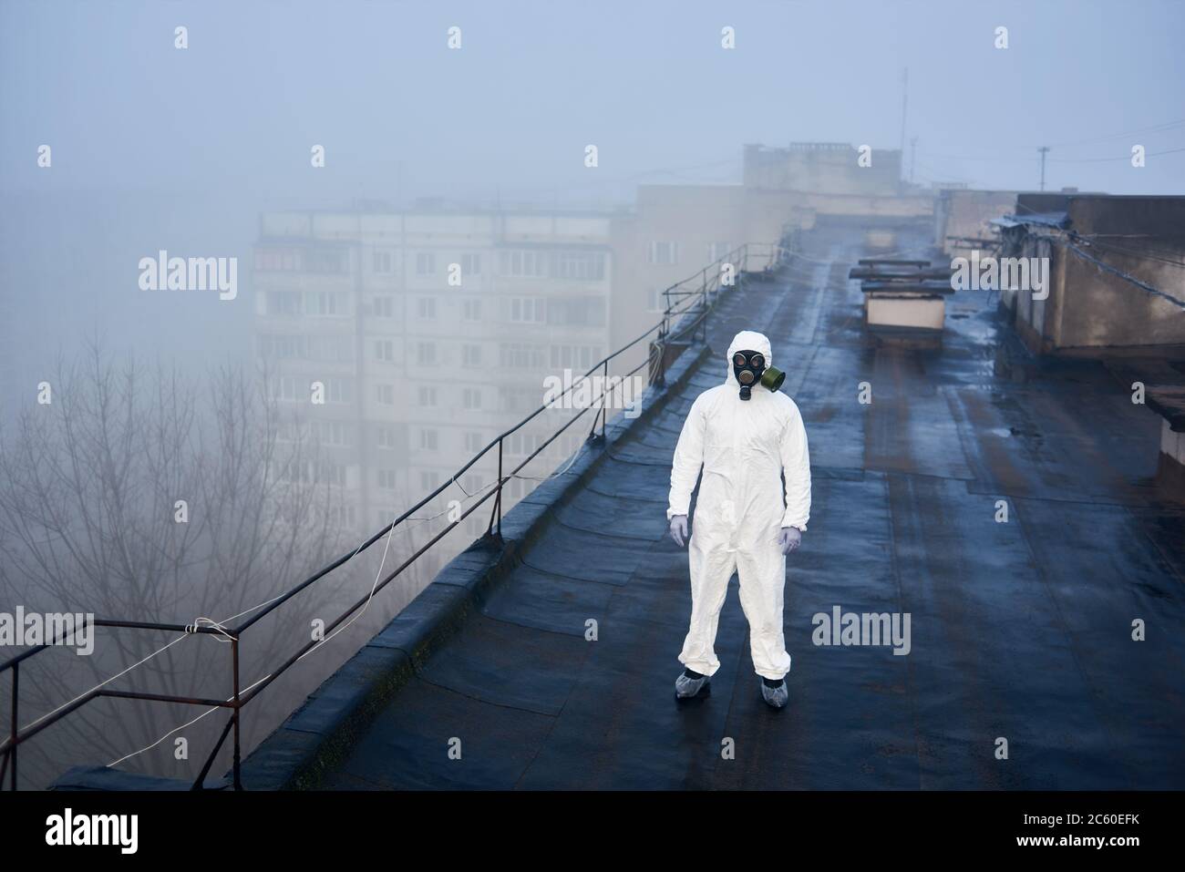 Uomo scienziato in piedi sul tetto di blocco di appartamenti, indossando tuta di protezione bianca e maschera a gas, imparando l'ambiente per le ricerche future. Vista dall'alto Foto Stock