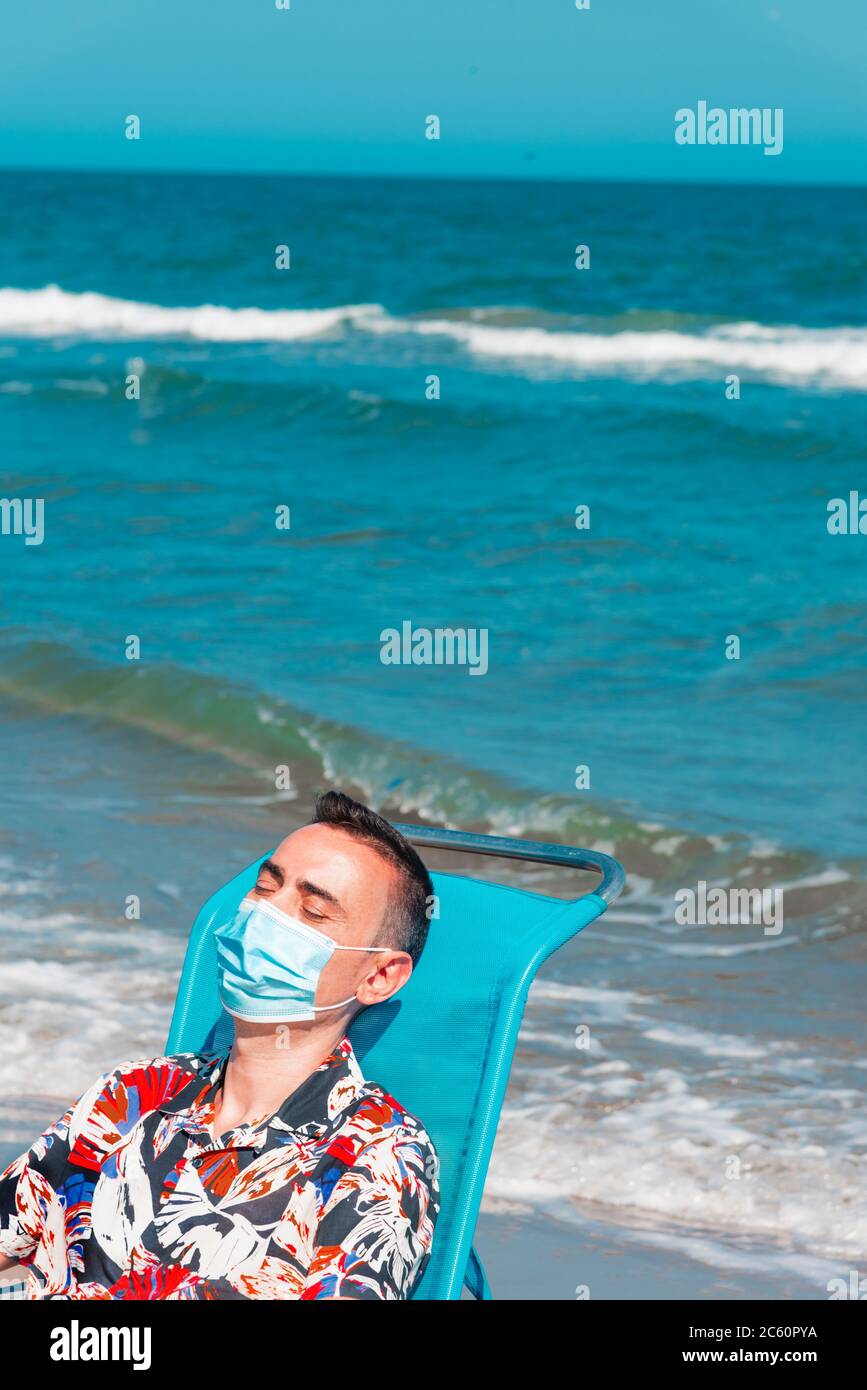primo piano di un uomo caucasico che indossa una maschera chirurgica blu mentre è seduto su una sedia a sdraio sulla spiaggia, con il mare sullo sfondo Foto Stock