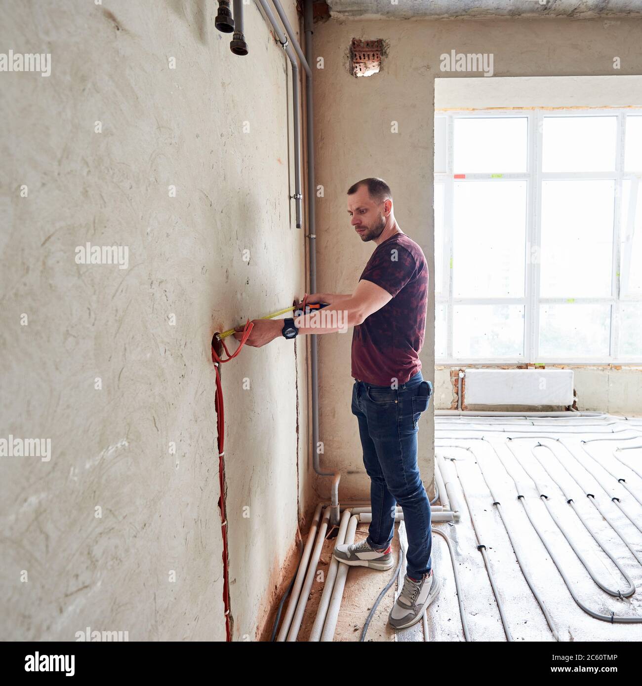 Giovane uomo in appartamento vuoto incompiuto che misura la parete con  l'aiuto di un metro a nastro, in piedi su pavimento riscaldamento sistema  di tubi contro finestra panoramica. Concetto di rinnovo e