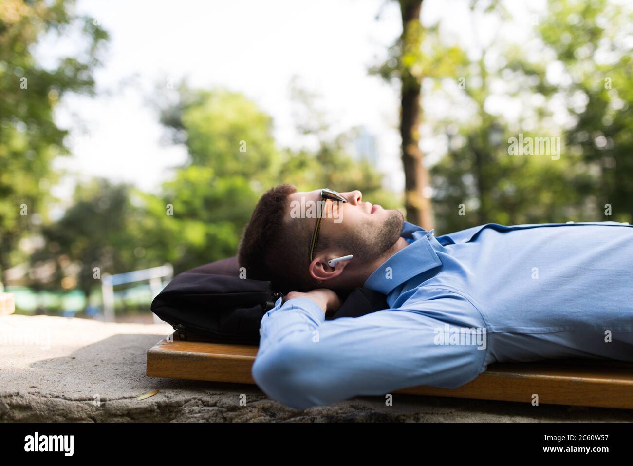 Giovane uomo bello in camicia blu e occhiali da sole con auricolari  wireless sognare dormire sulla panca nel parco della città Foto stock -  Alamy