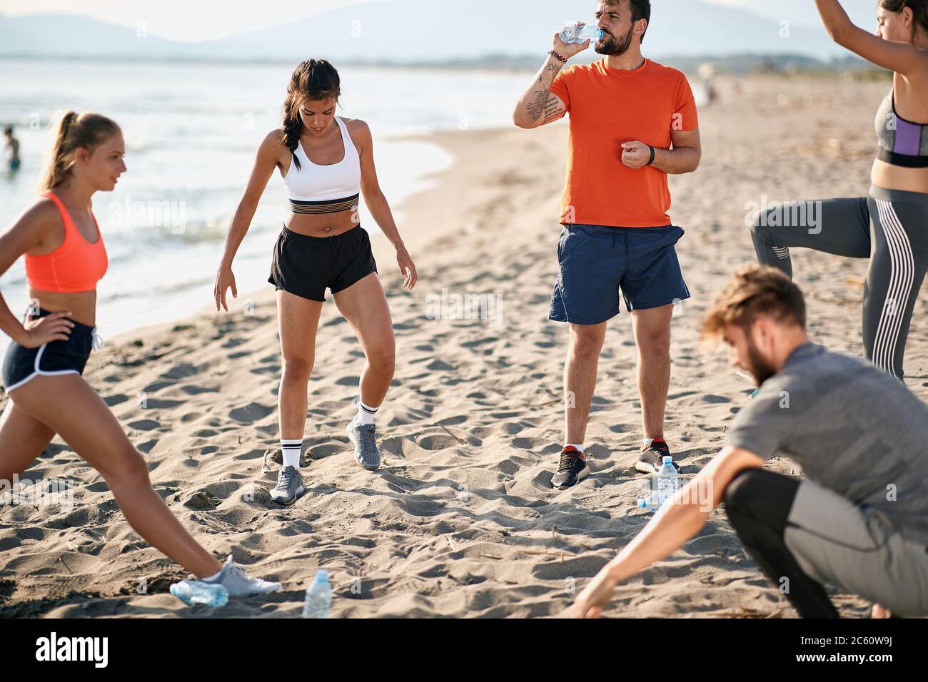 gruppo di giovani adulti caucasici che fanno esercizi sulla spiaggia sabbiosa. ragazzo ardito bere acqua da una bottiglia di plastica Foto Stock