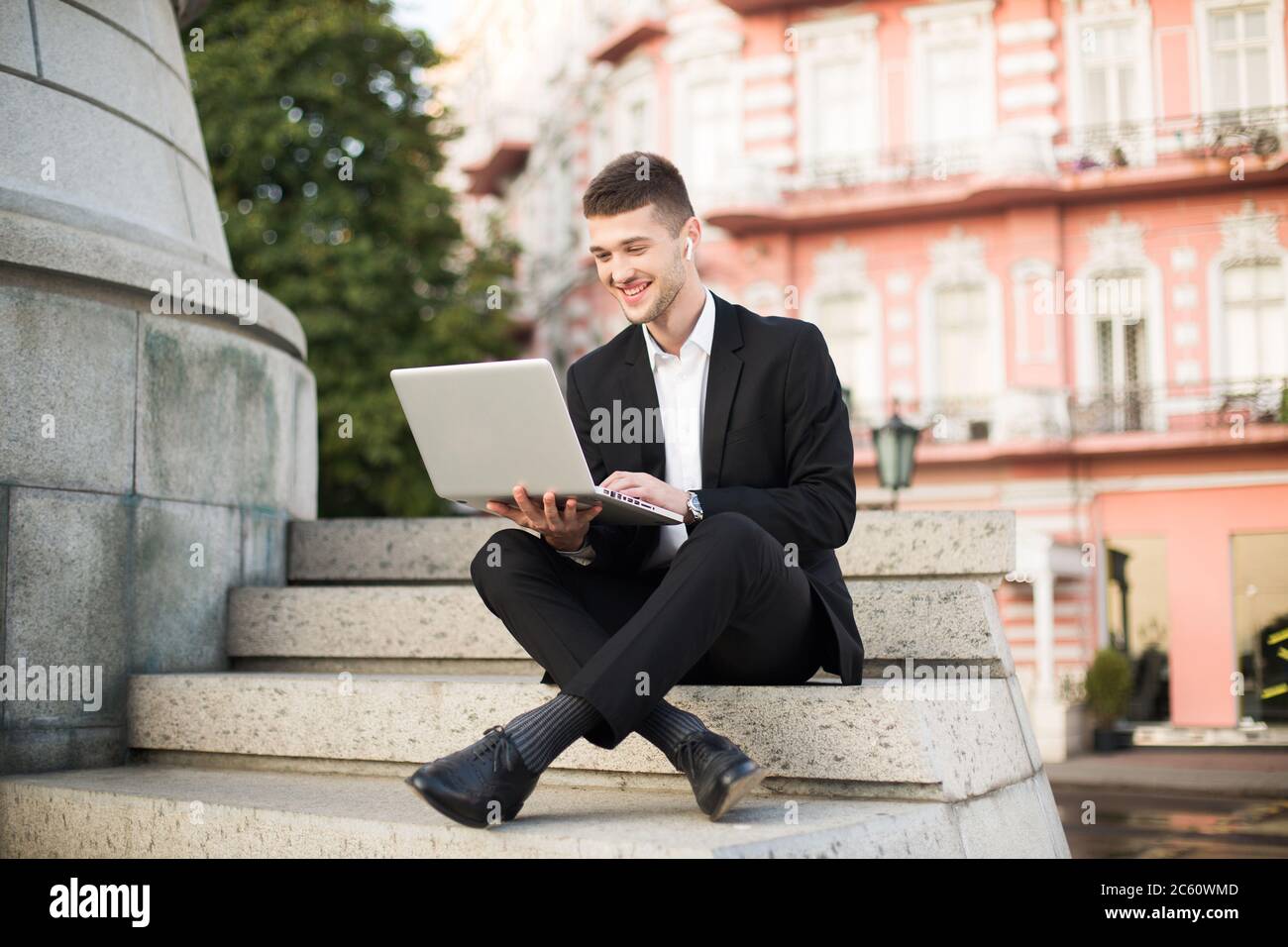 Giovane gioioso uomo d'affari in classico abito nero con auricolari wireless che lavora felicemente con il computer portatile seduto su piccole scale mentre si passa il tempo Foto Stock