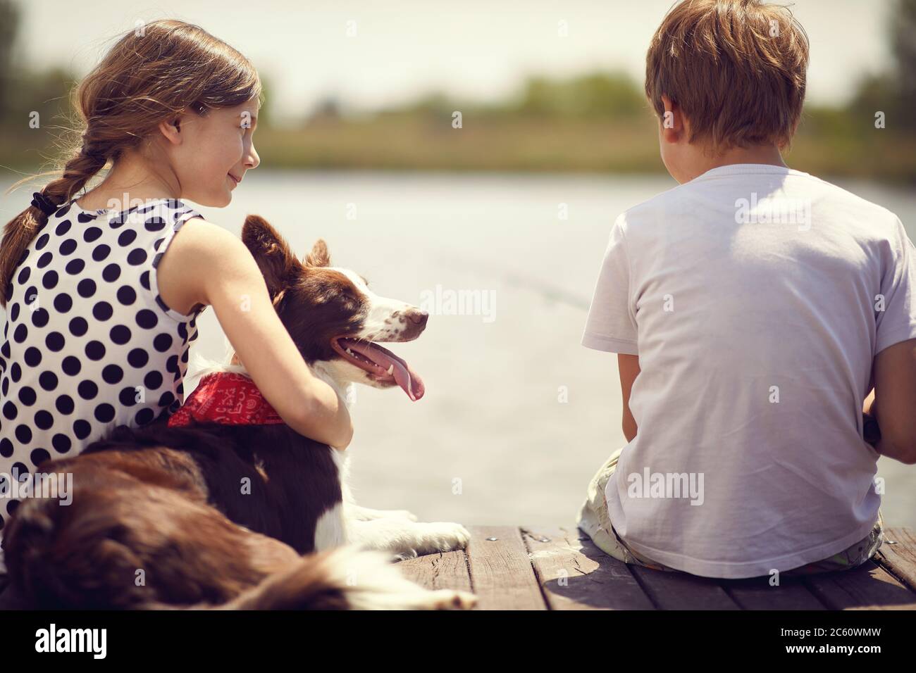 Ragazzino e ragazza con il cane seduto su un molo di legno e la pesca in uno stagno. Foto Stock