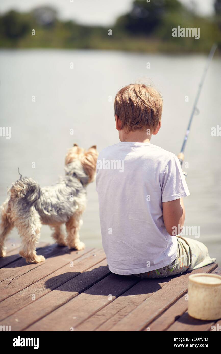 Ragazzo piccolo con il cane seduto su un molo di legno e la pesca in uno stagno. Foto Stock