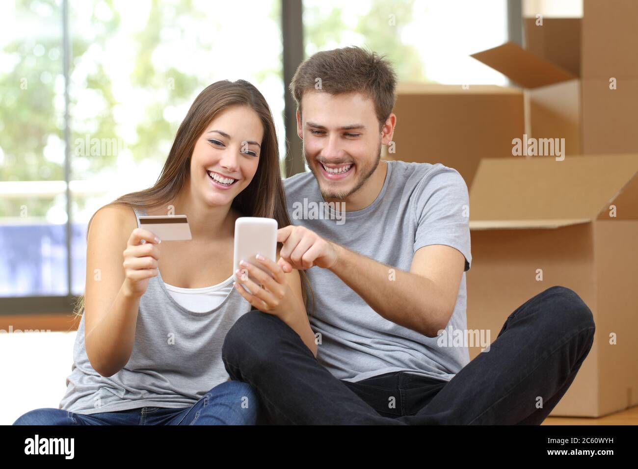 Coppia felice che paga in linea su smartphone con la carta di credito che si muove a casa seduta sul pavimento circondato da scatole Foto Stock
