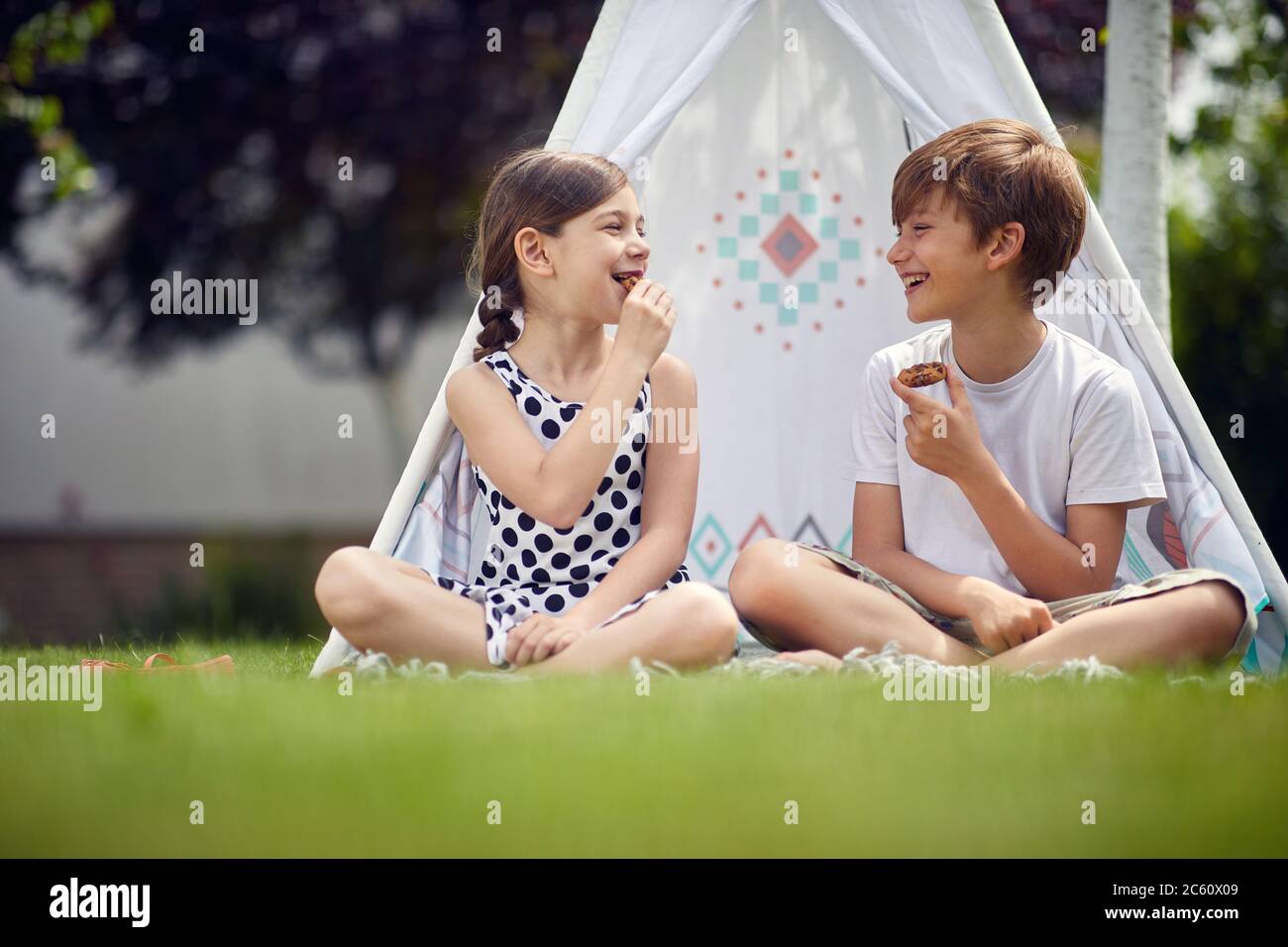 Divertimento estivo. Sorridente ragazzo e ragazza che gioca in cortile in teepee e mangia biscotti. Foto Stock