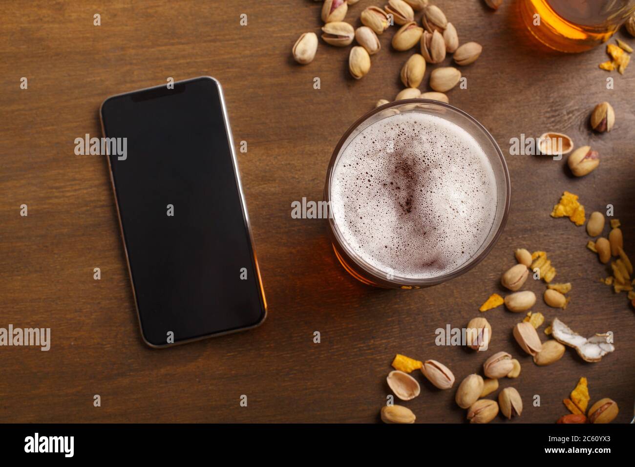 Birra e tecnologia. Smartphone e ale con schiuma su vetro, pistacchio e patatine sparse Foto Stock