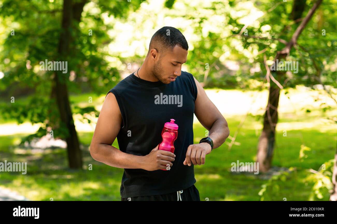 Corridore afroamericano con bottiglia d'acqua che controlla il suo smartwatch dopo l'allenamento al parco Foto Stock