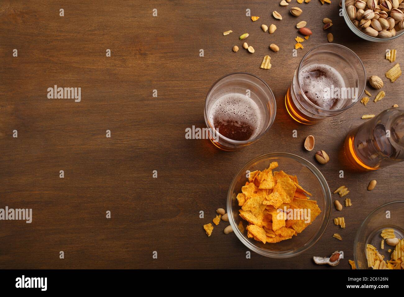 Due bicchieri di birra e bottiglia incompiuta, pistacchi, patatine e nachos in lastre di vetro e sparsi Foto Stock