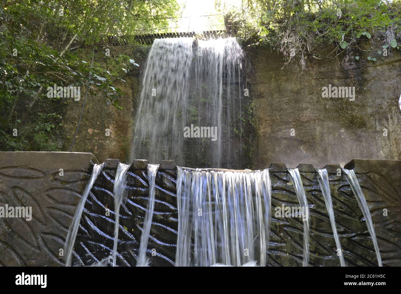 Cascata. Cascata costruita in cemento con acqua in caduta, vegetazione e alberi in una vista dal basso verso l'alto, con disegni di pesci nel calcestruzzo Foto Stock