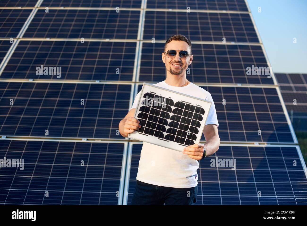 Sorridente giovane uomo che tiene un piccolo modulo fv guardando la telecamera, impianto solare dietro la schiena, concetto di energia alternativa Foto Stock