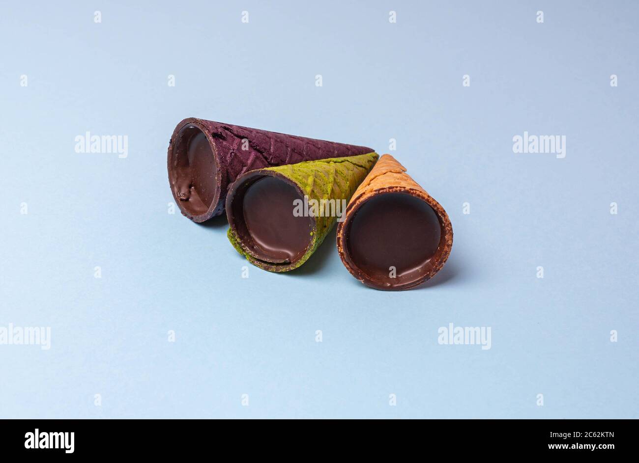 Corna di cialda viola, marrone e verde in glassa di cioccolato su sfondo blu chiaro, piatto. Dessert con waffle, primo piano, spazio per fotocopie Foto Stock