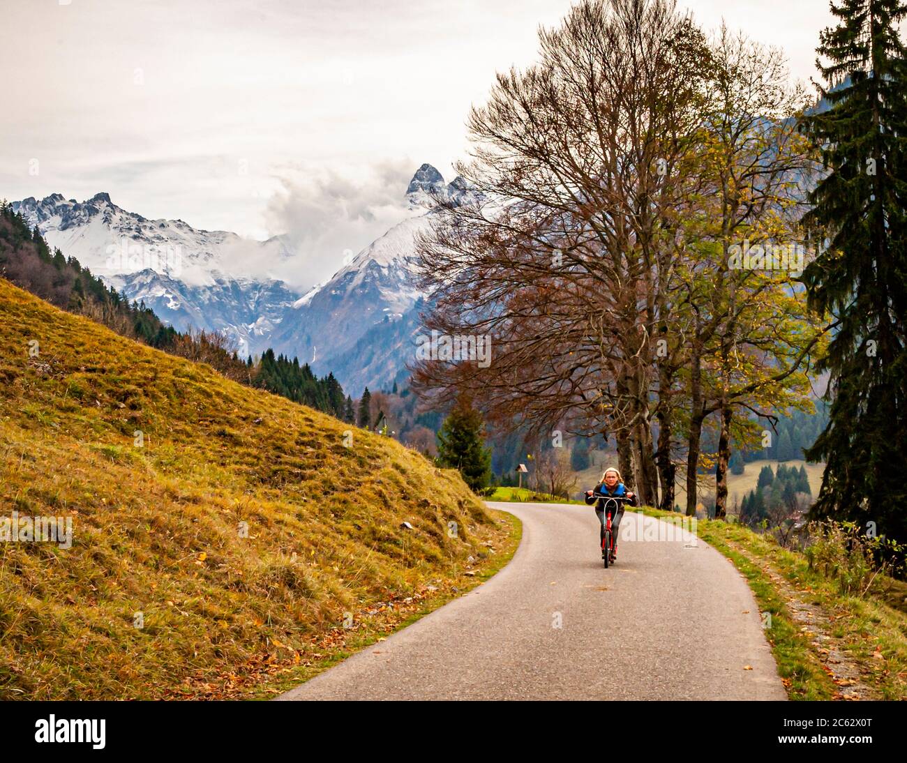 La pattinatrice di kick-scooter Angela Berg percorre una strada vicino all'Oy-Alm presso Oberstdorf, Bavaria, Germania, con le Alpi di Allgäu coperte di neve che si ergono alle sue spalle. Foto Stock