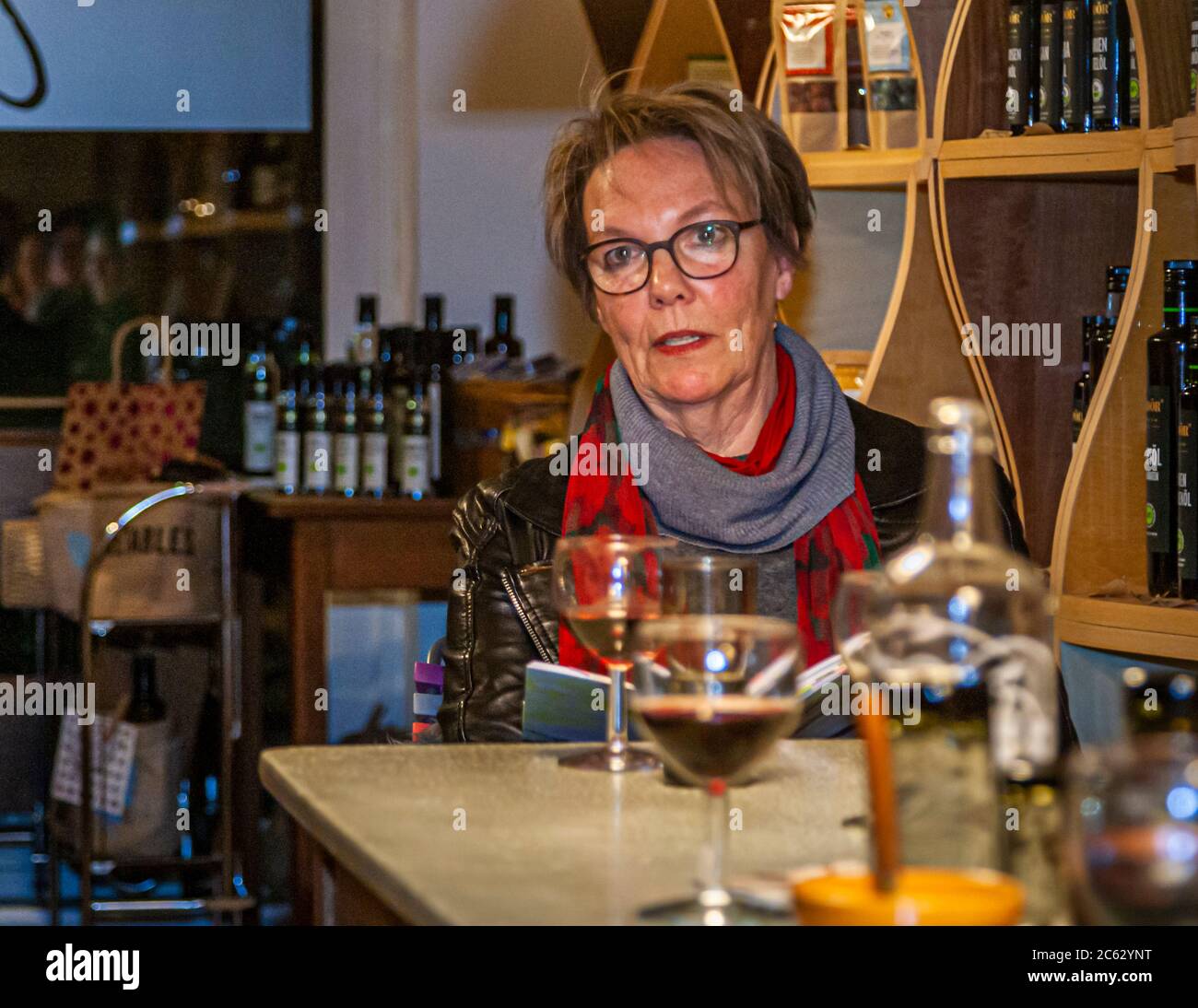 La scrittrice Janne Gronen siede con un libro aperto durante una lettura, al negozio di gastronomia "Peter sei selig – Genusspunkt Küche" a Grevenbroich, in Germania, con un bicchiere di vino rosso davanti a lei. Scaffali pieni di bottiglie di olio d'oliva riempiono lo sfondo. Foto Stock