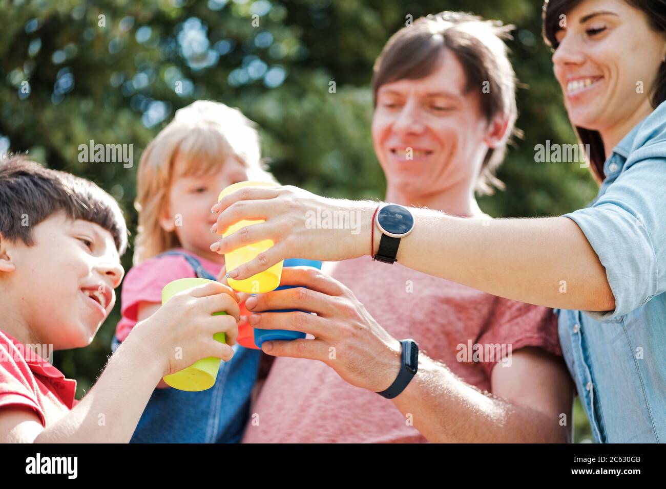 Una famiglia giovane e felice si tostano all'aperto con tazze colorate in un angolo basso, con vista ravvicinata dei volti sorridenti e messa a fuoco sulle mani Foto Stock
