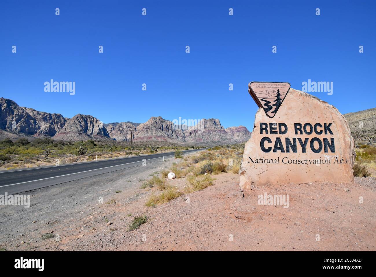 Una strada nel deserto passa accanto alle montagne con una striscia rossa e un cartello per la Red Rock Canyon National Conservation Area in una giornata con cielo blu chiaro. Foto Stock