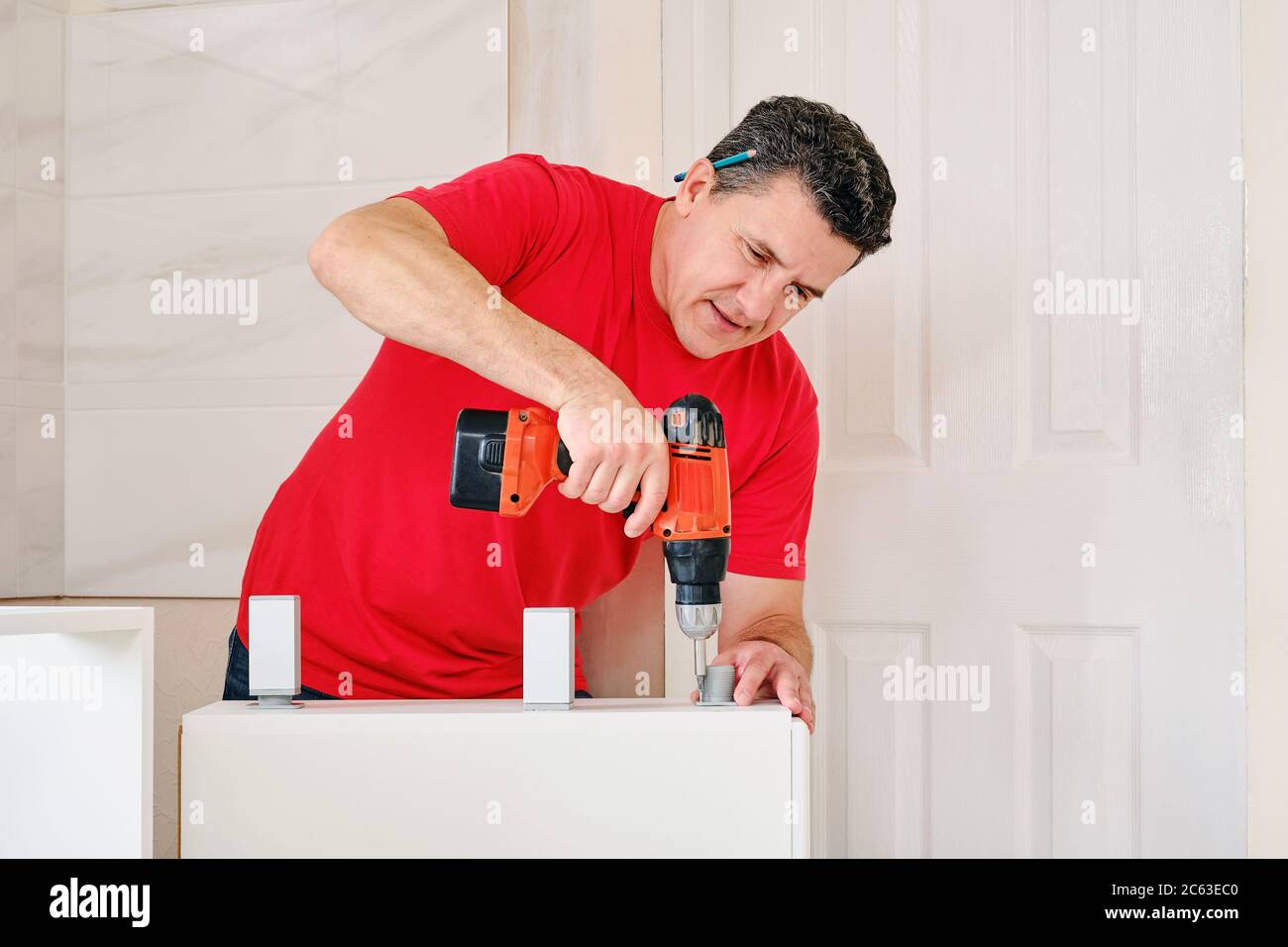 uomo bianco medio invecchiato avvitato le gambe del mobile in mobili di legno con avvitatore a batteria closeup. montaggio cucina in legno per la casa Foto Stock