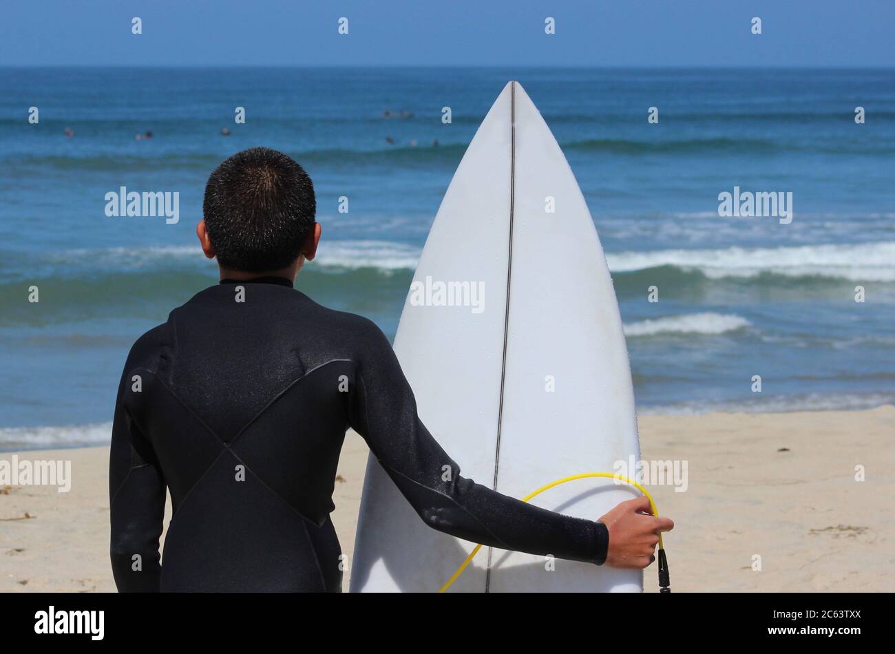 Un giovane surfista ispanico maschio si erge sulla spiaggia che tiene la sua tavola da surf e guarda le onde Foto Stock