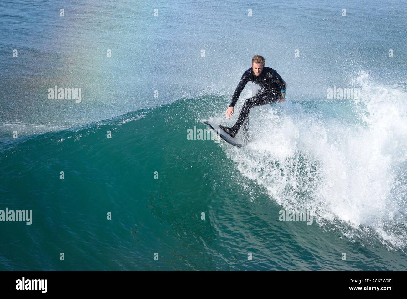 Giovane surfista maschile che cavalca una grande onda, un arcobaleno che si forma nella nebbia Foto Stock