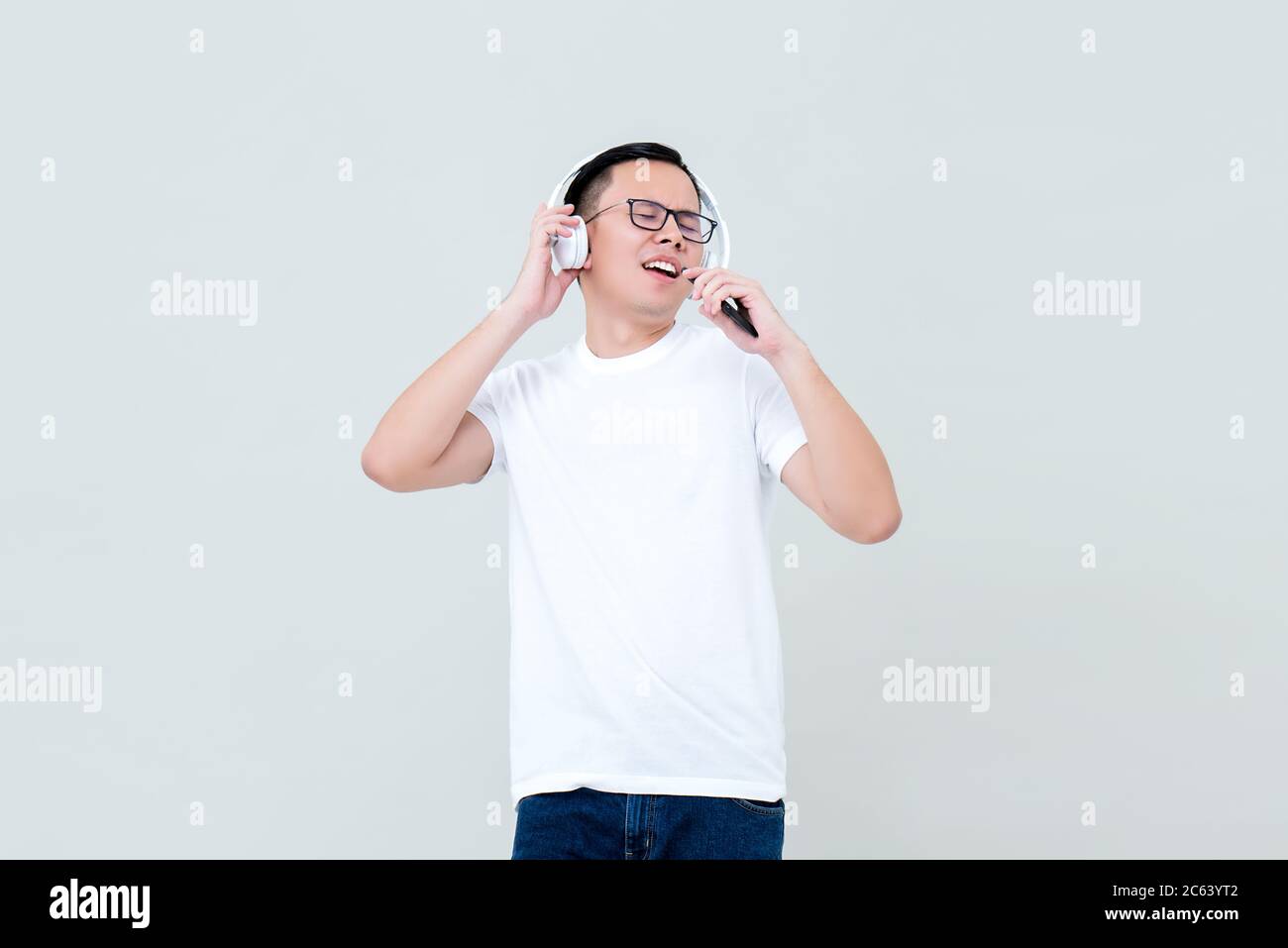 Giovane uomo asiatico felice che ascolta la musica dalle cuffie e canta isolato su sfondo grigio chiaro Foto Stock