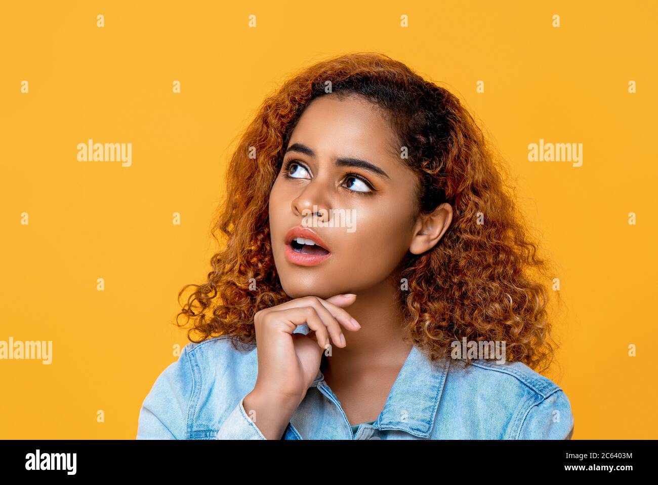Misto africano donna pensare con la mano sul mento e gli occhi guardando fino a spazio da parte isolato su sfondo giallo Foto Stock