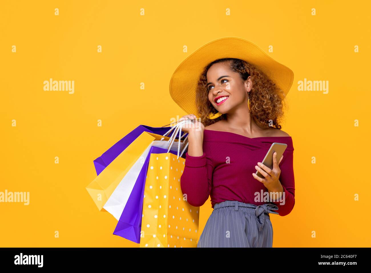 Donna di razza mista sorridente alla moda con borse colorate e telefono cellulare isolato su sfondo giallo per la vendita estiva conept Foto Stock