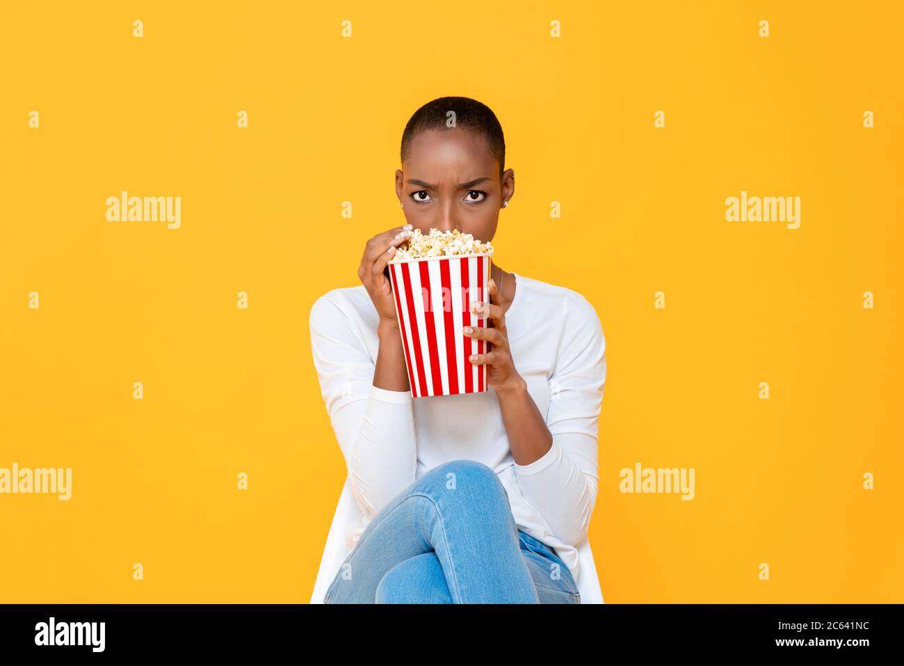 Giovane donna afro-americana spaventata guardando film horror e mangiare popcorn su sfondo giallo isolato Foto Stock