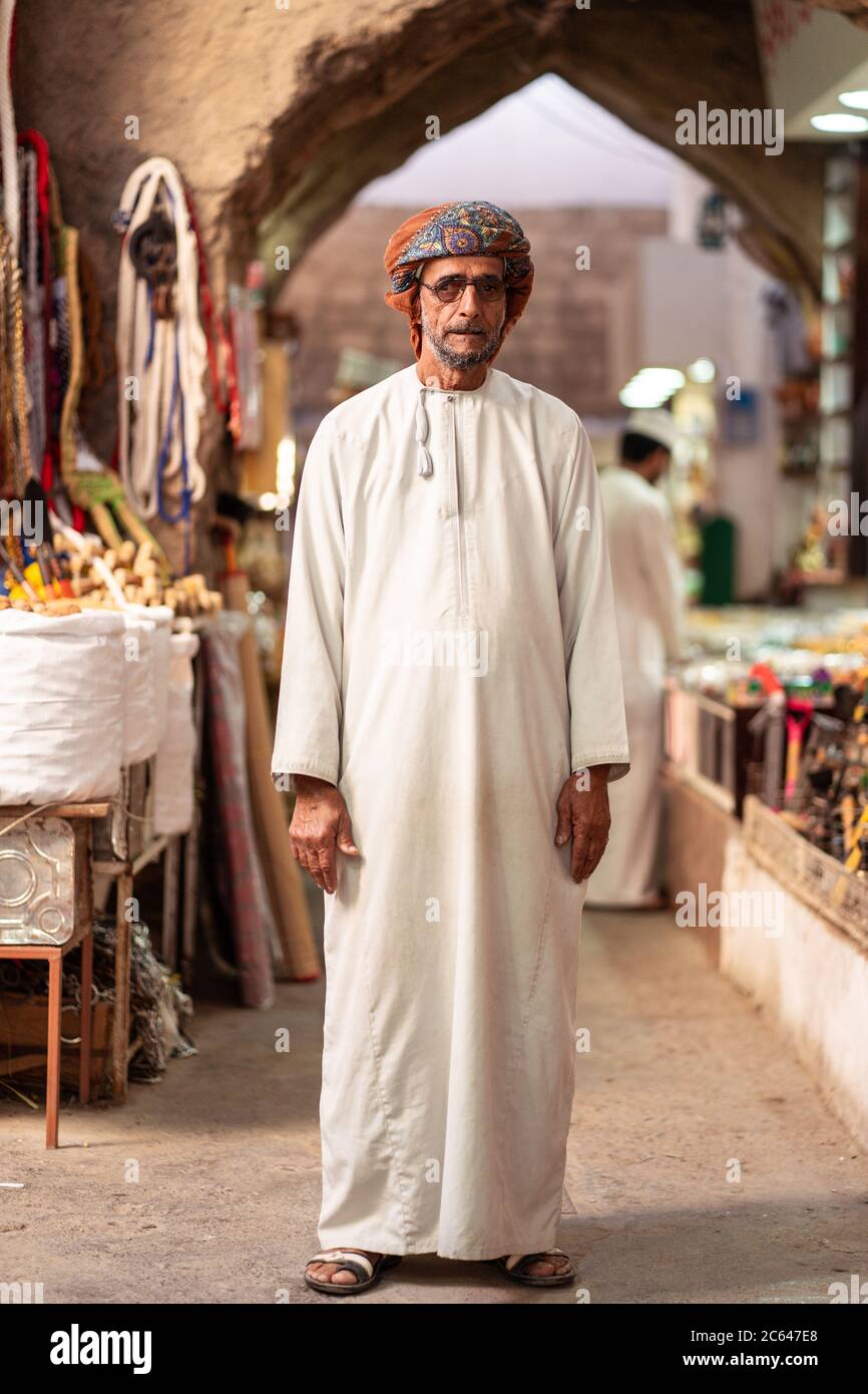 Nizwa / Oman - 15 febbraio 2020: Ritratto di vecchio uomo musulmano Omani con abiti tradizionali bianchi in piedi nel suq coperto di Nizwa con cancello ad arco Foto Stock