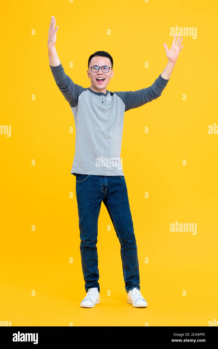 Ritratto a corpo pieno di allegro giovane asiatico che solleva entrambe le braccia in aria isolato su sfondo giallo Foto Stock