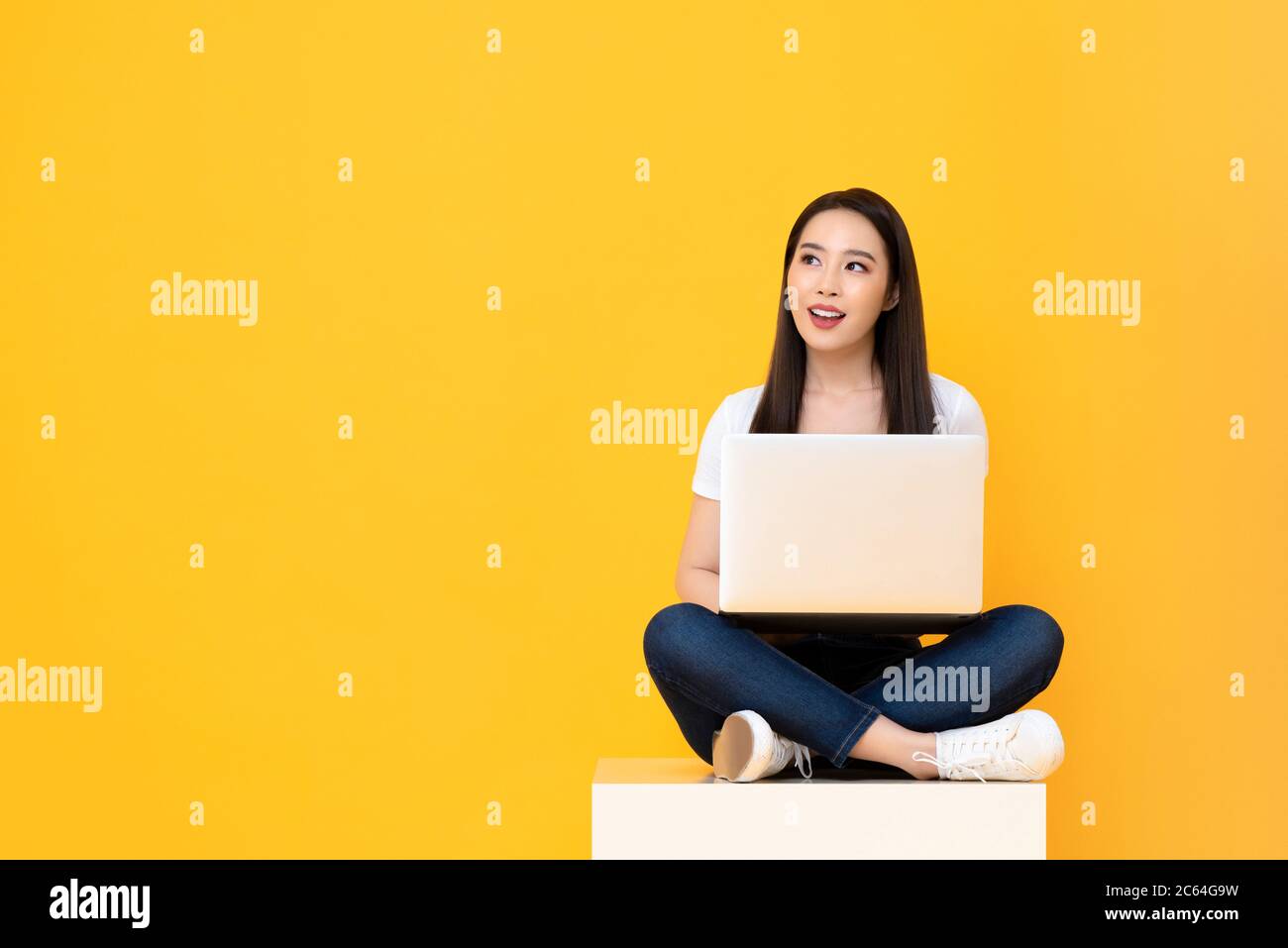 Felice attraente giovane donna asiatica con computer portatile seduto a gambe incrociate e guardando spazio vuoto su sfondo giallo studio Foto Stock