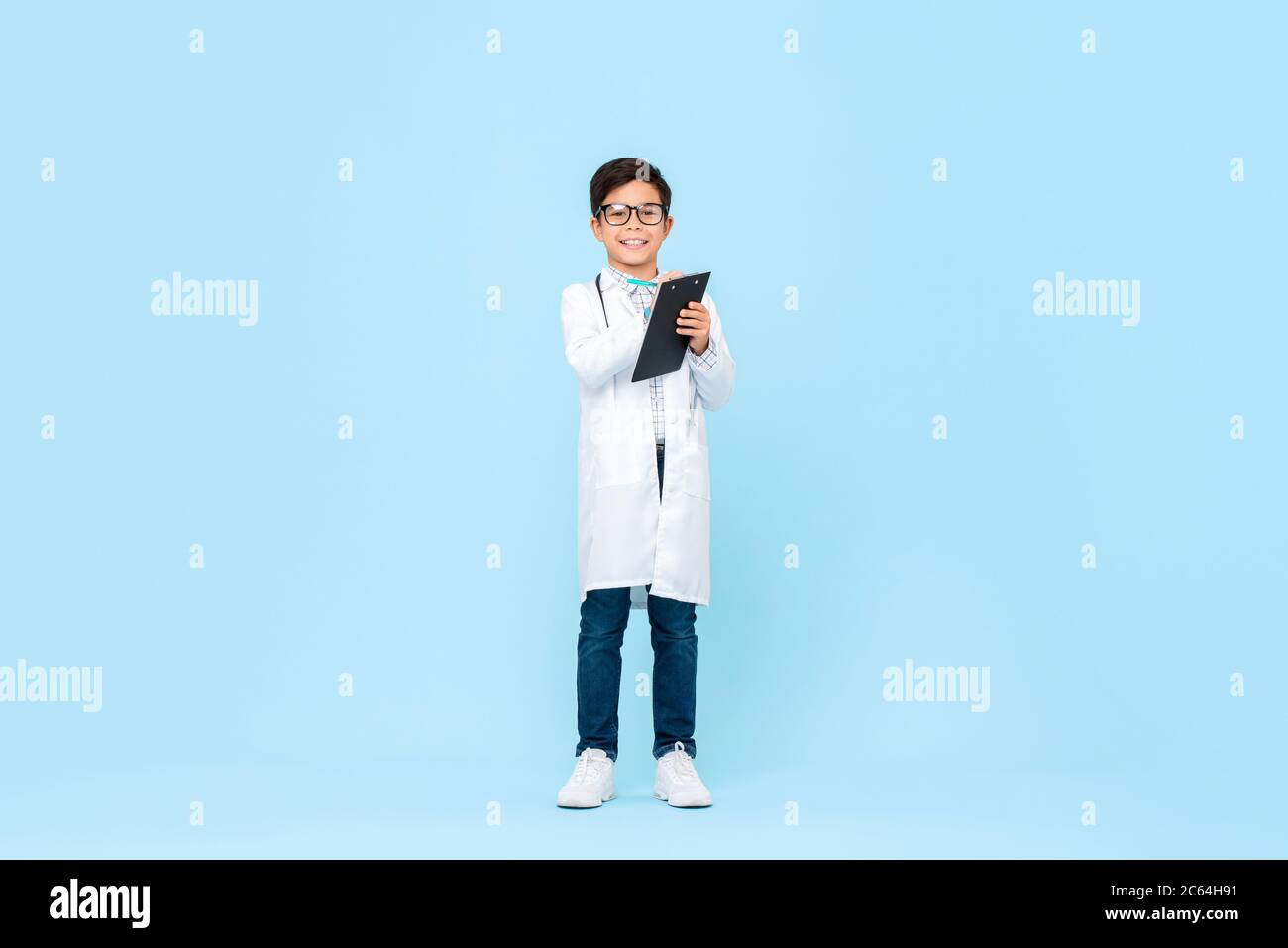 Ritratto di lunghezza intera di bambino asiatico sorridente che aspira ad essere medico futuro scrittura prescrizione negli appunti in blu sfondo isolato Foto Stock