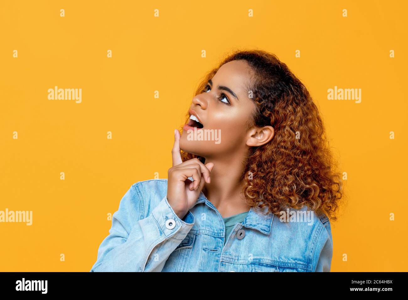 Primo piano ritratto vista laterale della giovane curiosa bella donna afroamericana guardando in alto mentre tocca il mento con un dito in giallo studio isolato Foto Stock
