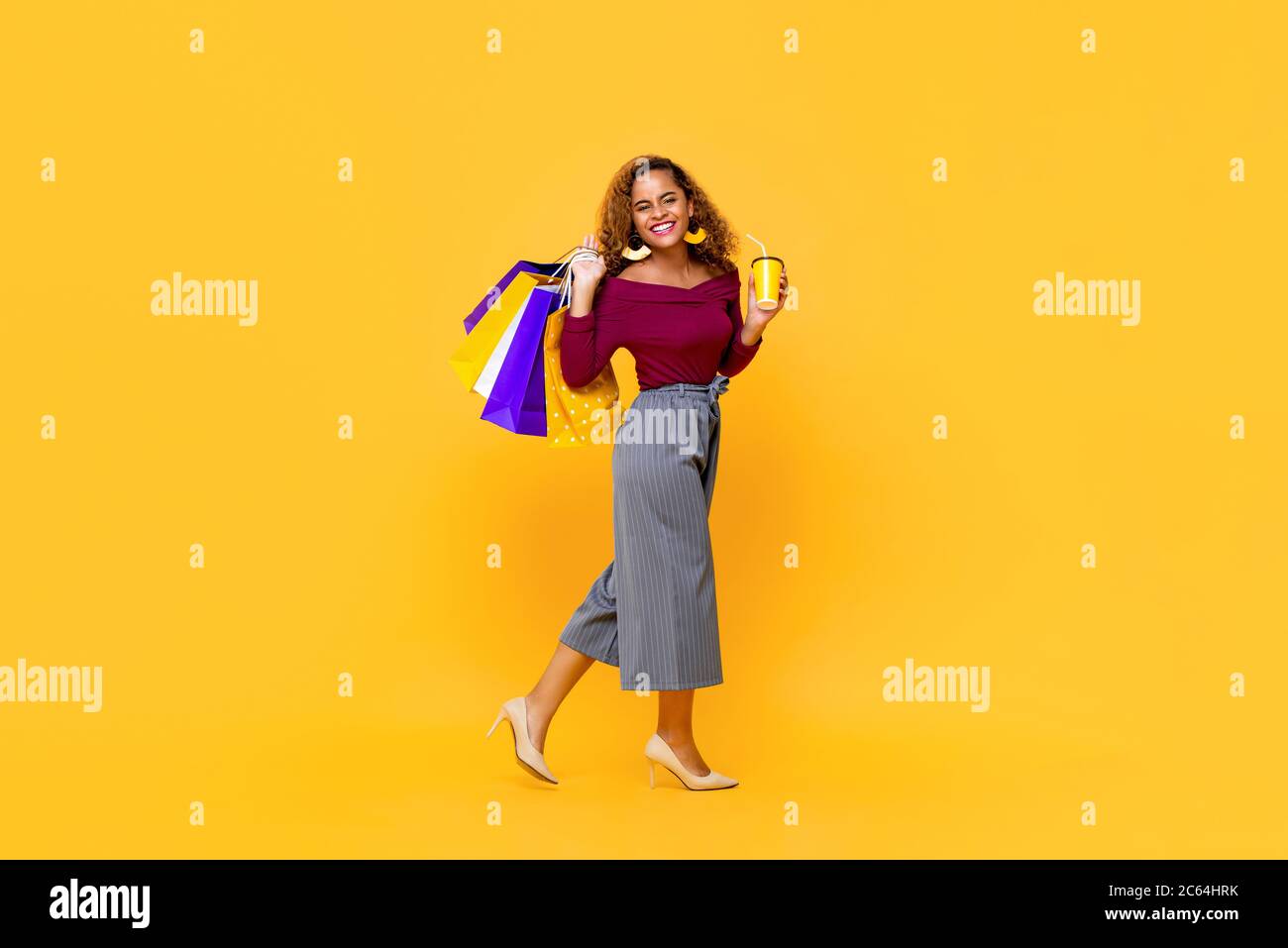 Ritratto a lunghezza intera di giovane allegra donna afro-americana sorridente che cammina mentre tiene borse per lo shopping e tazza di bevanda in studio isolato Foto Stock