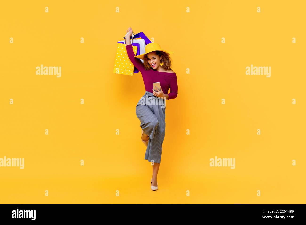 Divertente ritratto a lunghezza intera di felice giovane attraente shopaholic afroamericano donna alzando borse per la spesa e tenendo il telefono cellulare in studio isolato Foto Stock