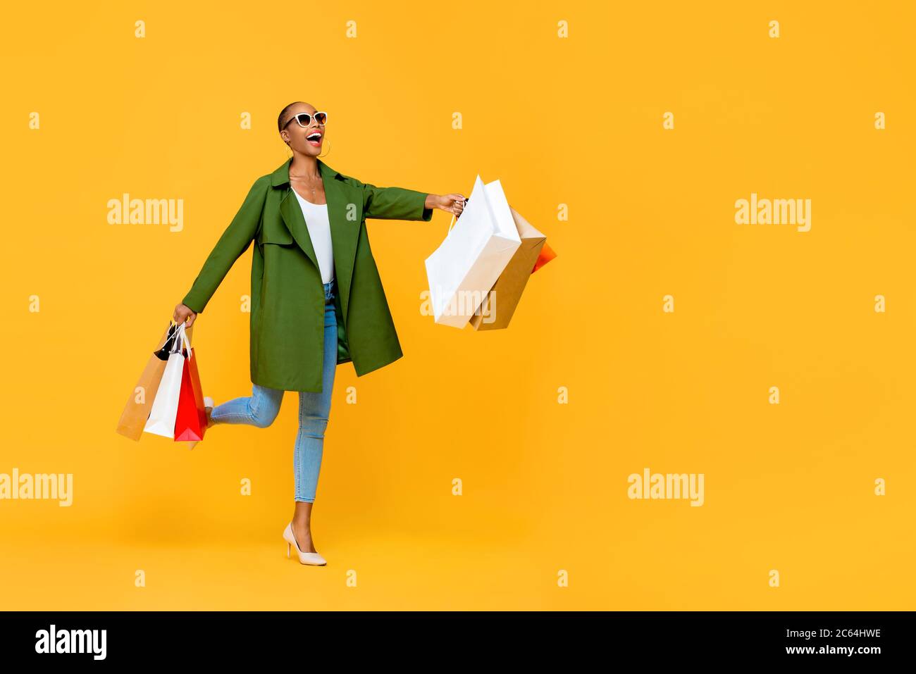 Ritratto a lunghezza intera di allegra attraente donna afroamericana che tiene borse per la spesa mentre si panzica in uno studio isolato sfondo giallo Foto Stock