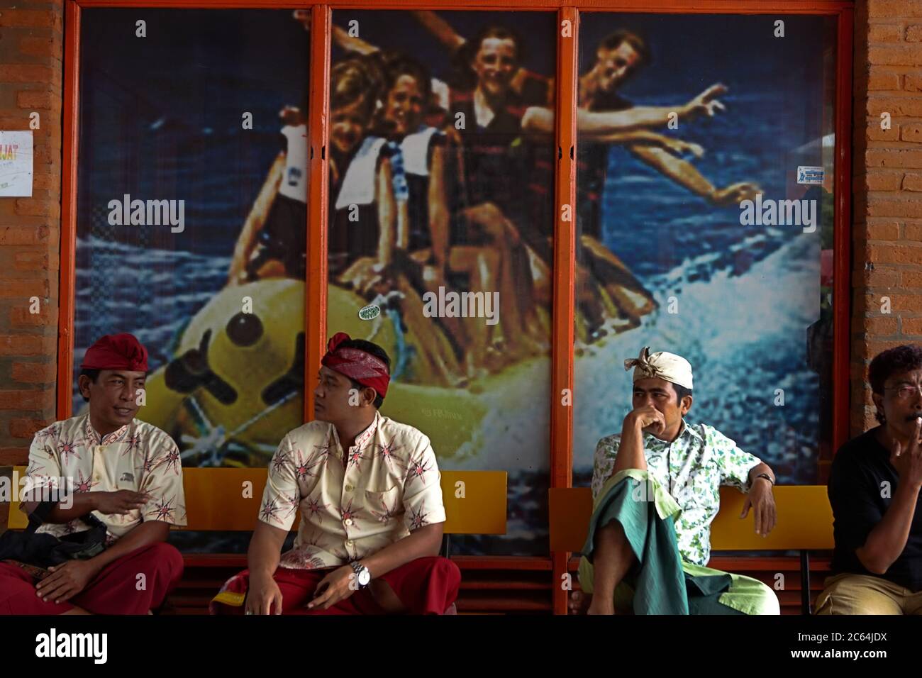 Uomini in abiti tradizionali balinesi che hanno una conversazione di fronte a un grande poster di sport acquatici a Bedugul, Bali, Indonesia. Foto Stock