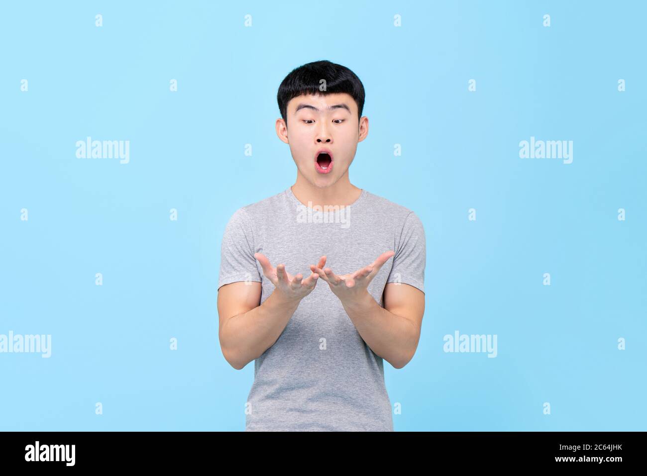 Divertente ritratto di un giovane asiatico stupito guardando entrambe le mani con bocca aperta in uno sfondo blu studio isolato Foto Stock