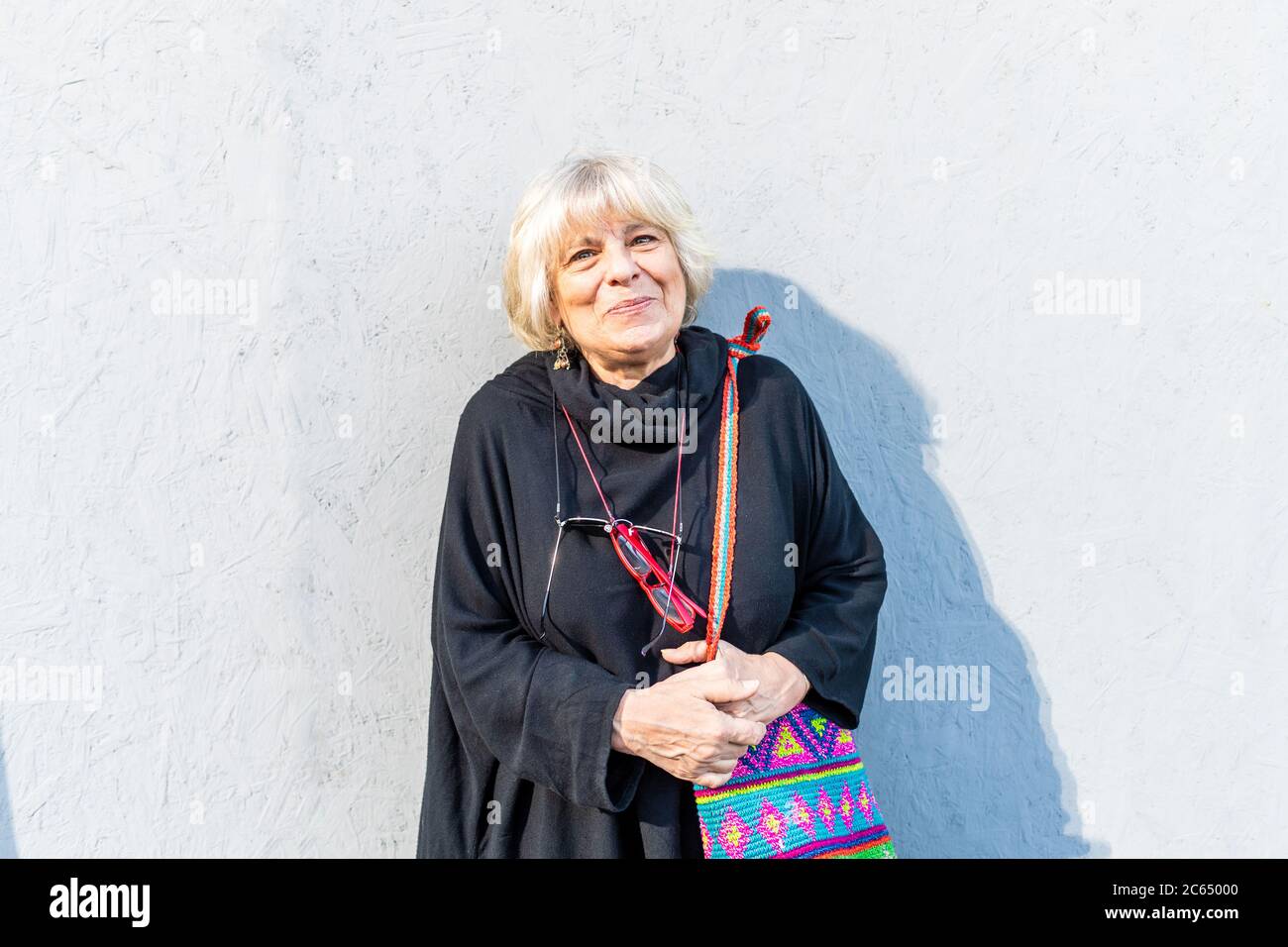 Ritratto di donna anziana con capelli biondi che indossa la parte superiore nera, in piedi davanti alla parete bianca, sorridente alla macchina fotografica. Foto Stock