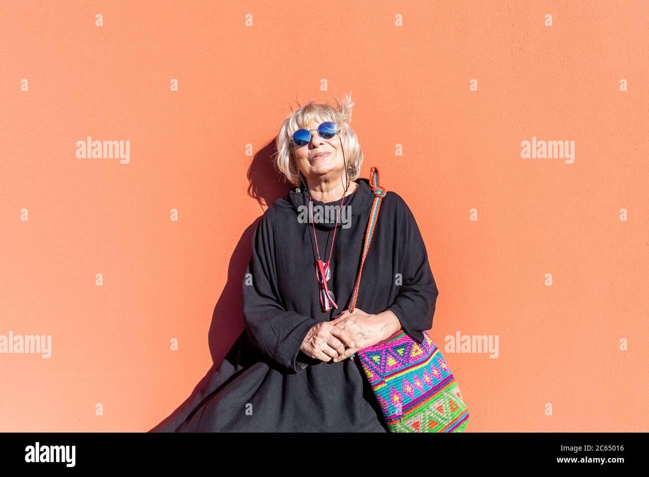 Ritratto di donna anziana con capelli biondi e occhiali da sole in piedi davanti alla parete arancione. Foto Stock