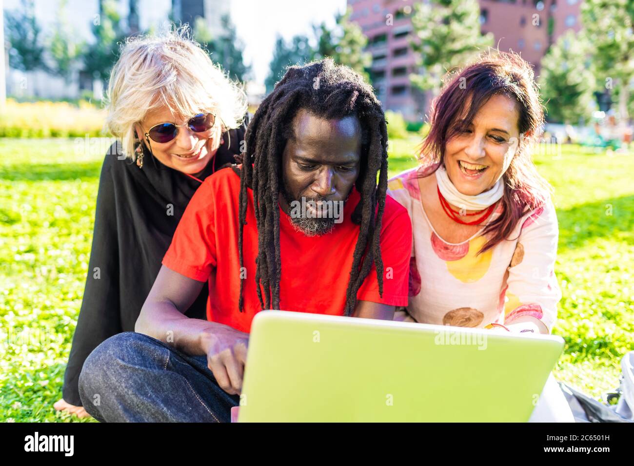 Uomo nero con dreadlock e due donne caucasiche seduti sul prato, guardando il computer portatile. Foto Stock